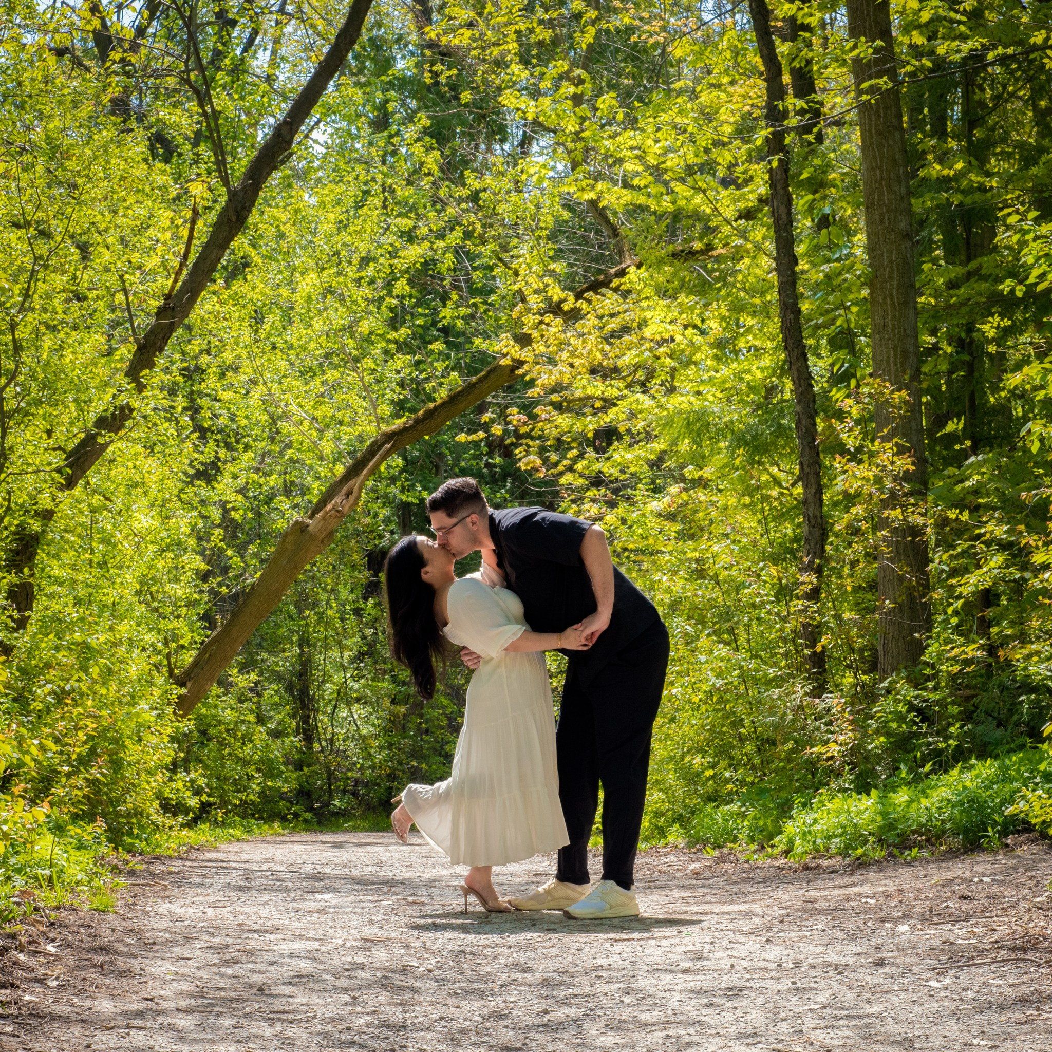 A couple mid-dip kiss on a sunlit gravel path lined with yellow-green trees