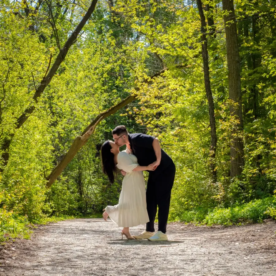 A couple mid-dip kiss on a sunlit gravel path lined with yellow-green trees