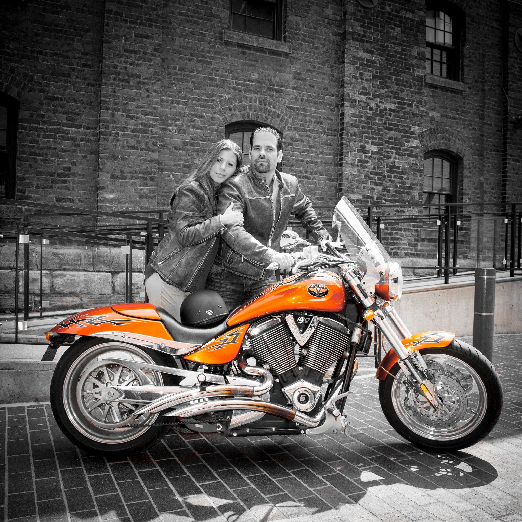 A couple seated on an orange motorcycle against a black-and-white brick streetscape