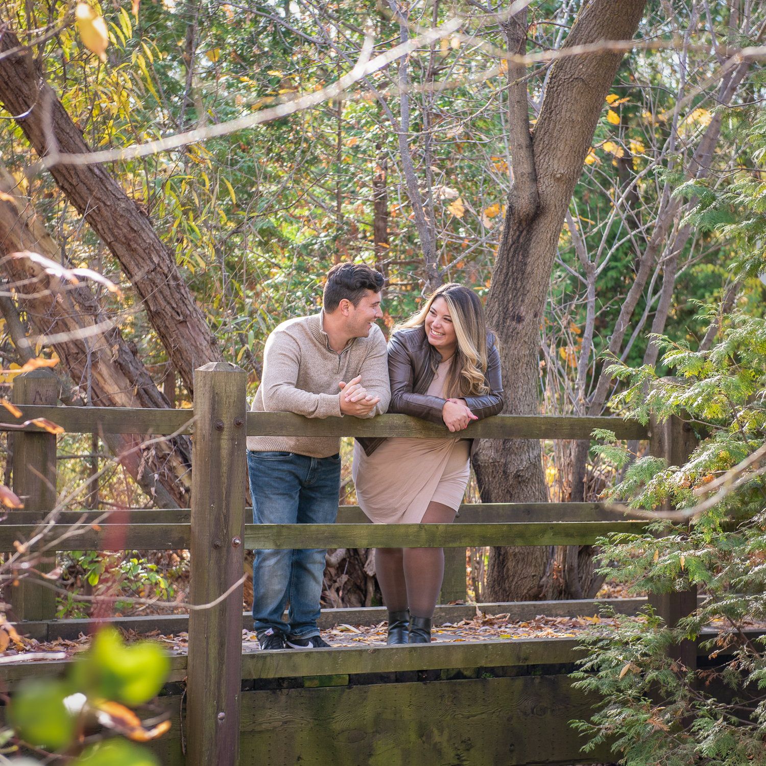 A couple leaning on a wooden footbridge railing with autumn willows behind them