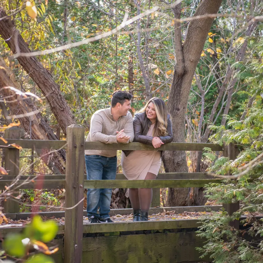 A couple leaning on a wooden footbridge railing with autumn willows behind them