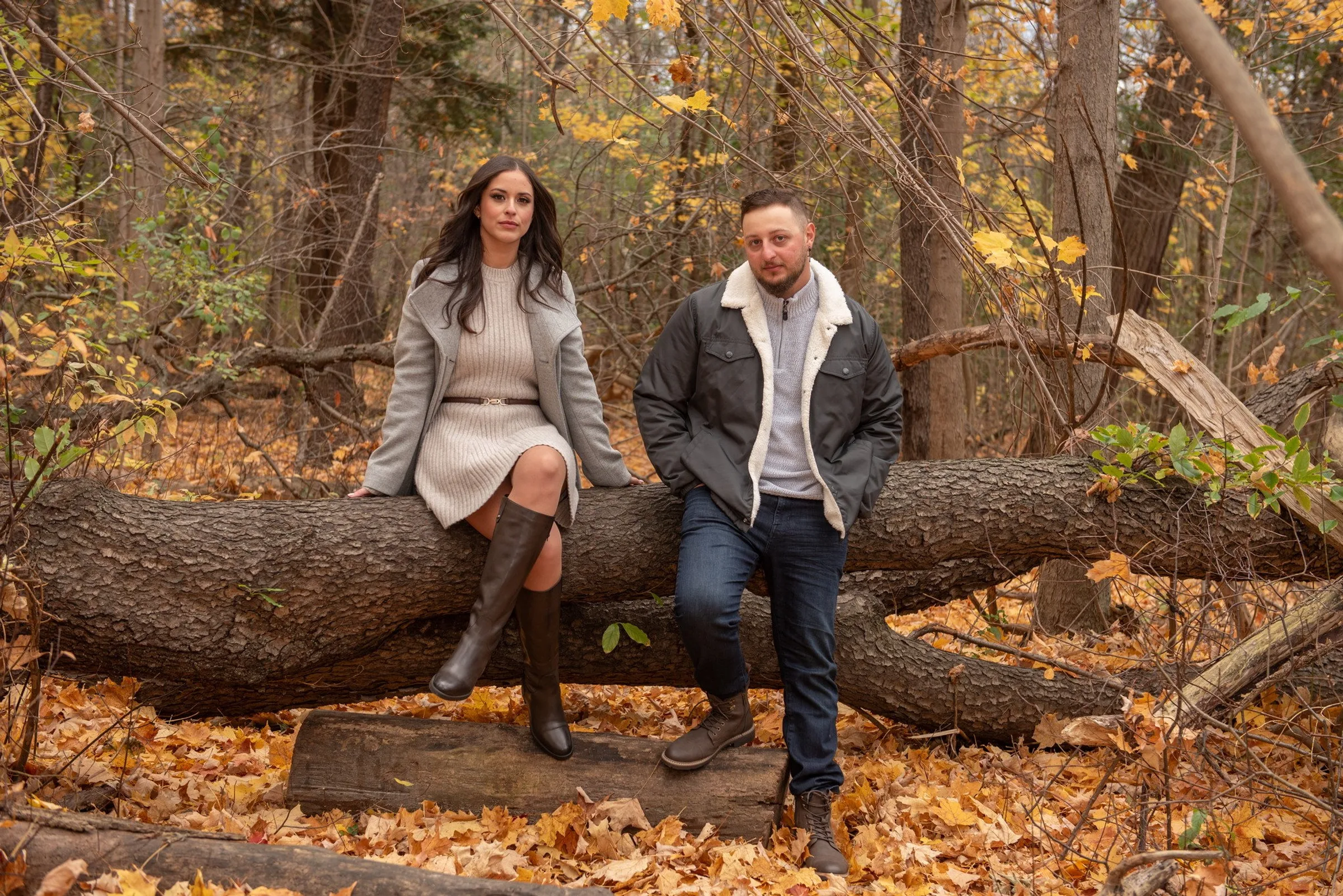 A couple in autumn coats sitting side by side on a fallen log, surrounded by yellow maple leaves
