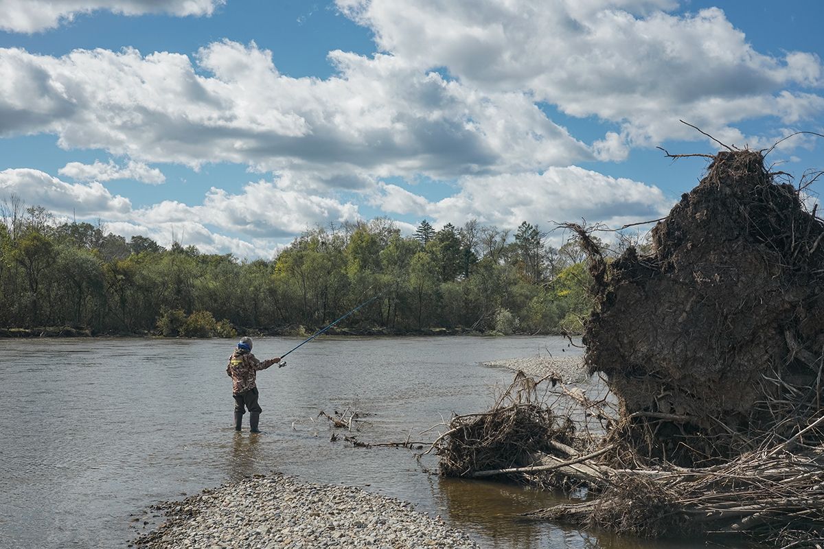 An Udege man stands in the Bikin River wearing fishing gear and holding a rod. Uprooted trees lie on the riverbank in the foreground; a forest stretches in the background. Clear day, Primorye.
