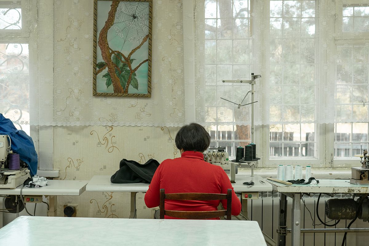 A woman in a red sweater sits at a sewing machine, working on a traditional garment. The room is decorated with lace curtains and a framed picture of a spiderweb. Bulava village, Khabarovsk Krai.