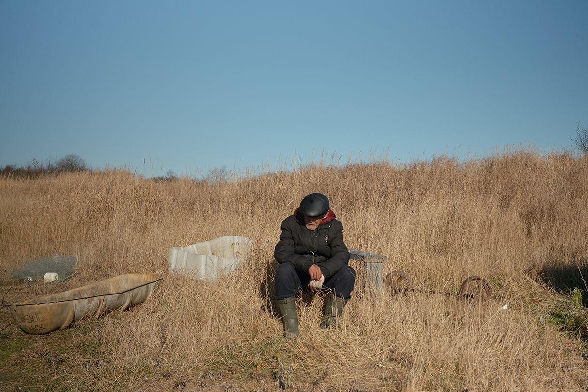 A man in a black jacket and rubber boots sits with his head lowered amid tall dry grass. Nearby are plastic containers and metal parts. Venskoye village, Sakhalin.