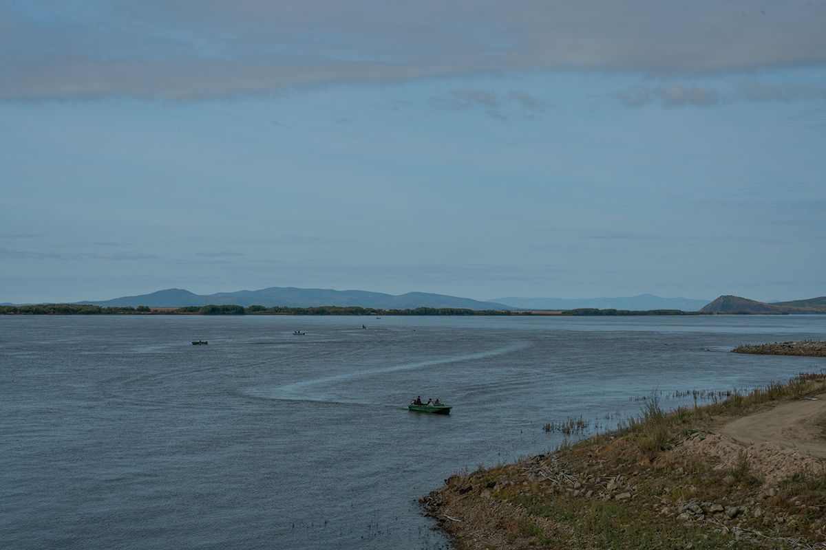 Several motorboats with fishers head out onto a wide river, with distant hills in the background. The shoreline on the right is grassy and rocky. Last day of the fishing season. Bulava village, Khabarovsk Krai.