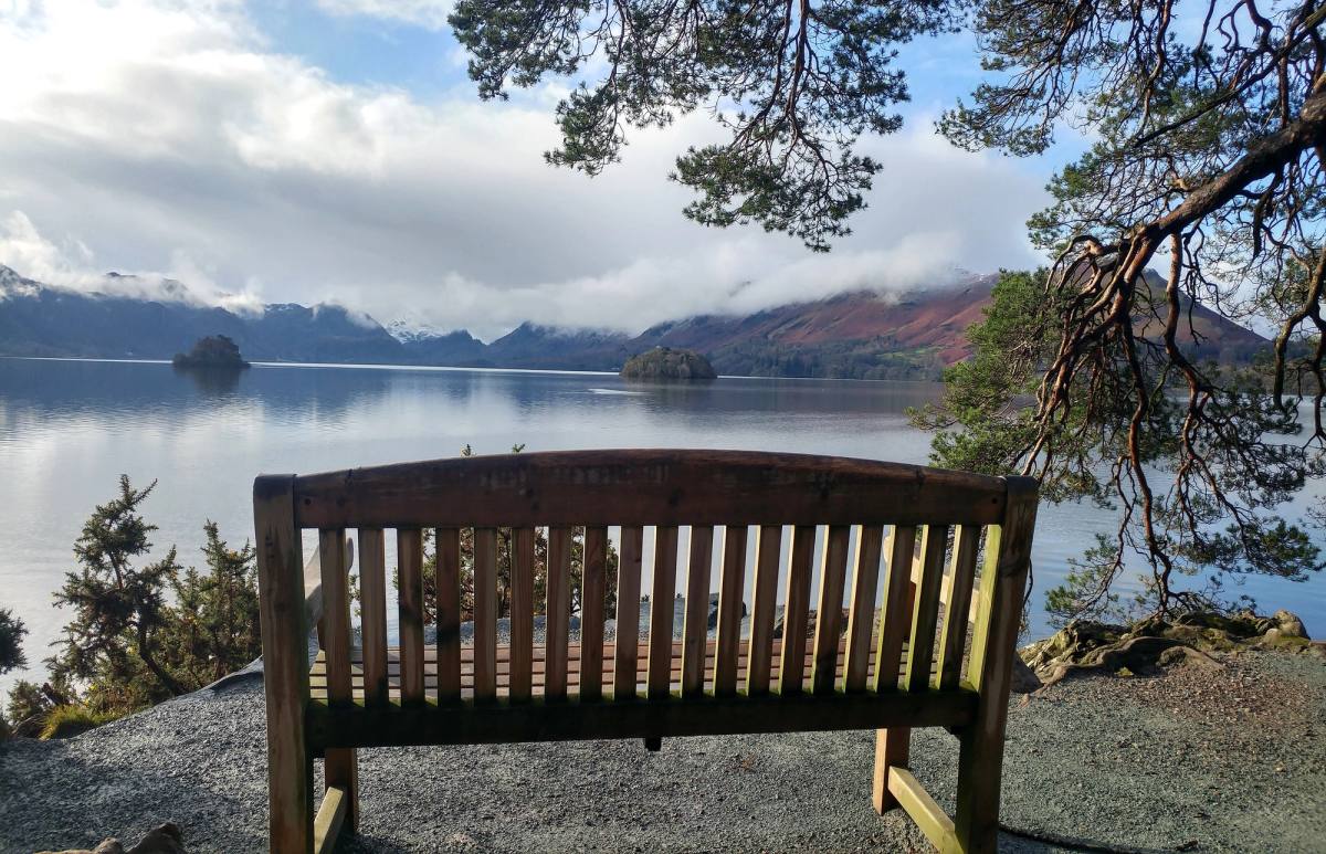 An empty wooden bench under trees looking out across Derwentwater to the fells