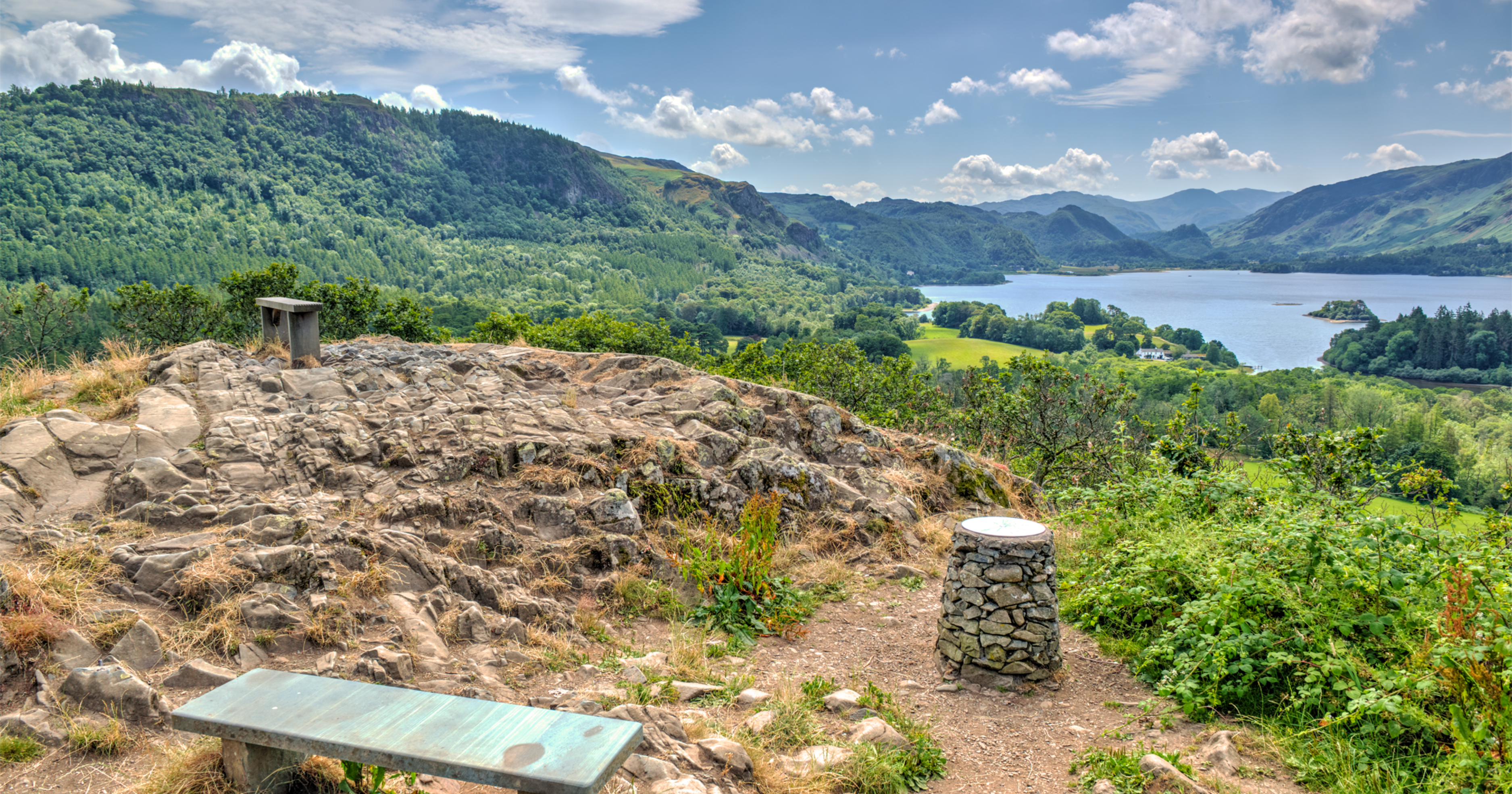 View from castlehead summit with a bench and a trig point waymarker with views across derwentwater