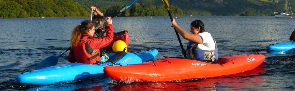 A family paddling two kayaks together on open water with the fells beyond