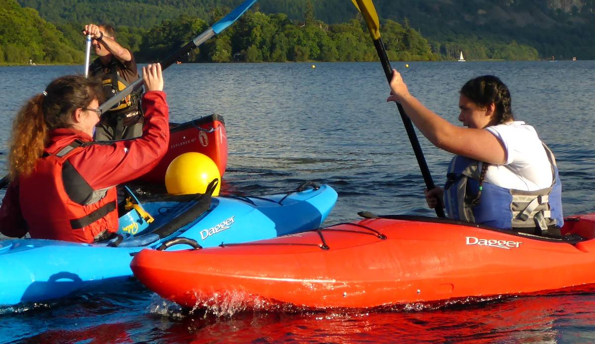 A family paddling two kayaks together on open water with the fells beyond