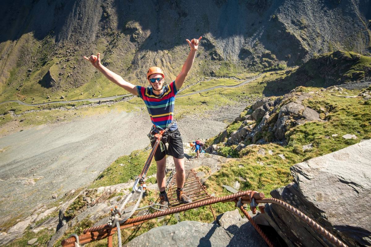 A climber on a via ferrata cable bridge with both arms raised in celebration