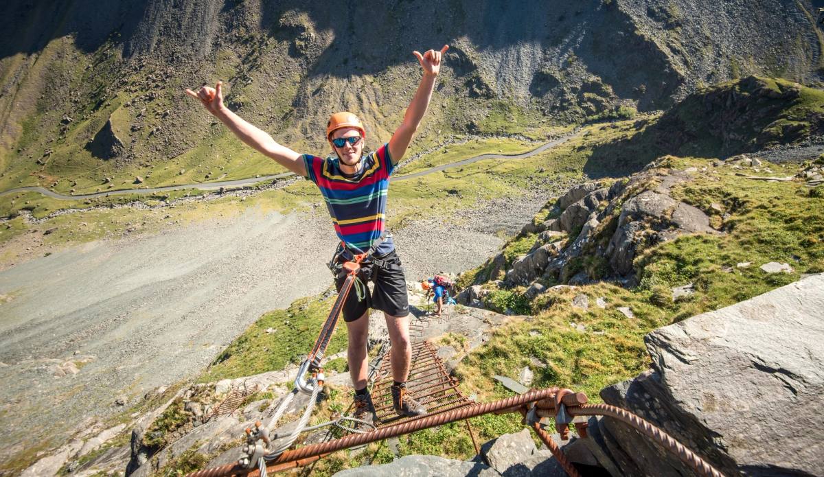 A climber on a via ferrata cable bridge with both arms raised in celebration