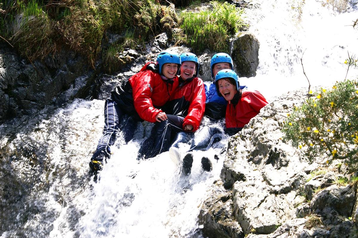 Four people in helmets and wetsuits sliding down a rushing waterfall