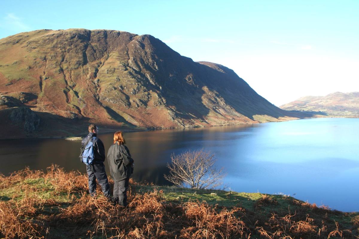 Two walkers on a bracken slope looking down over a lake to the fells opposite
