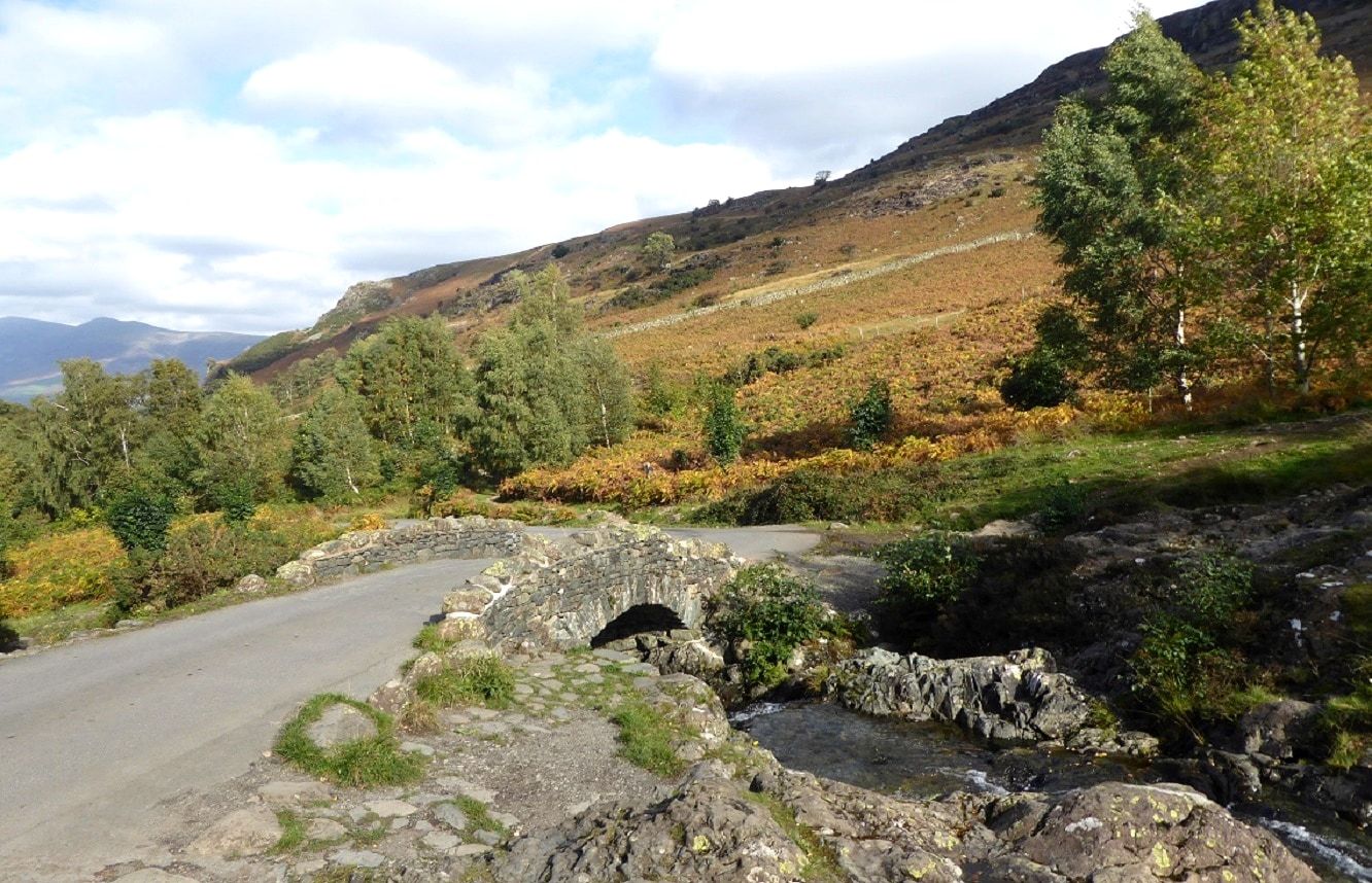 A low stone bridge carrying a narrow road across a beck below the fells