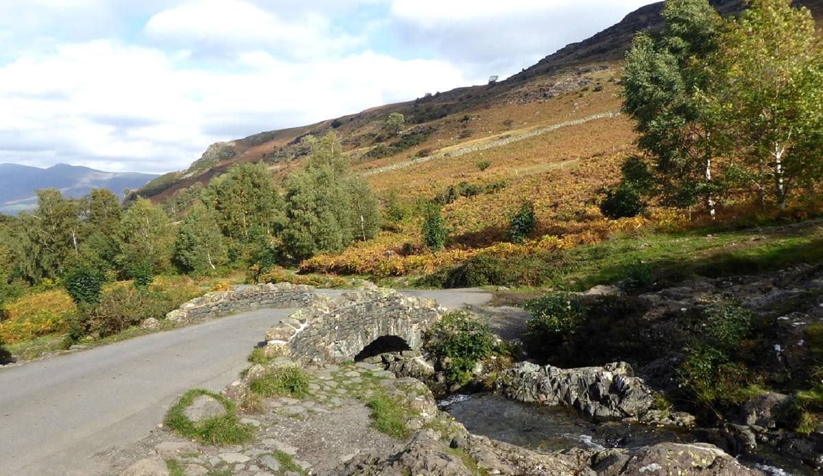 A low stone bridge carrying a narrow road across a beck below the fells