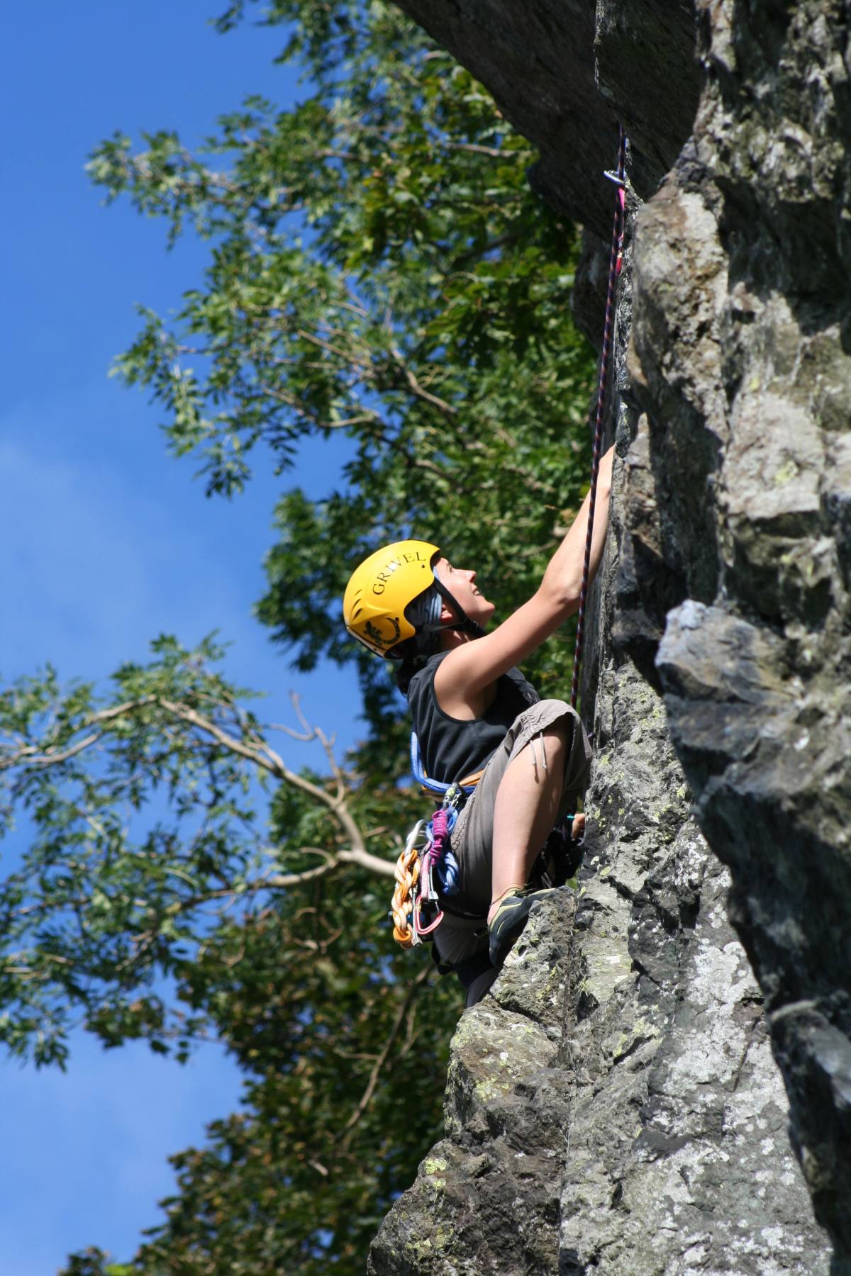 A climber in a yellow helmet on a crag, roped and reaching for the next hold