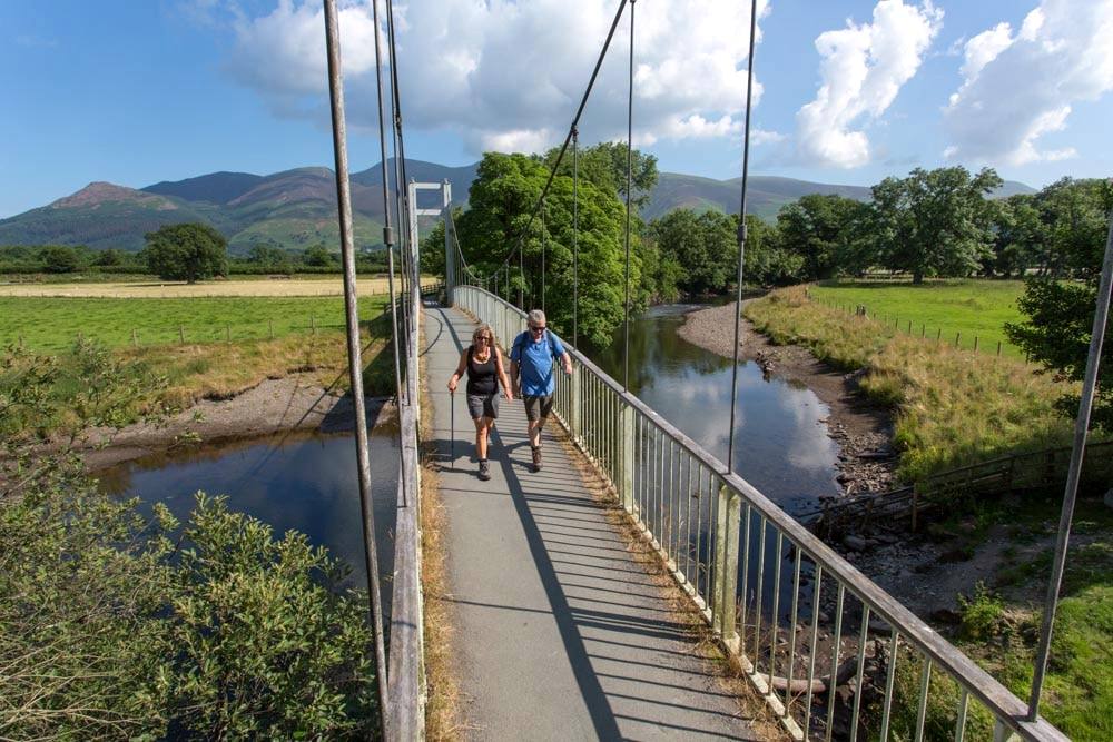 Two walkers crossing a suspension footbridge over the river with Skiddaw beyond