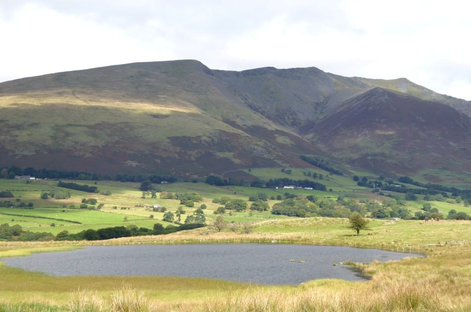 A shallow tarn in rough pasture with the long green ridge of Blencathra behind