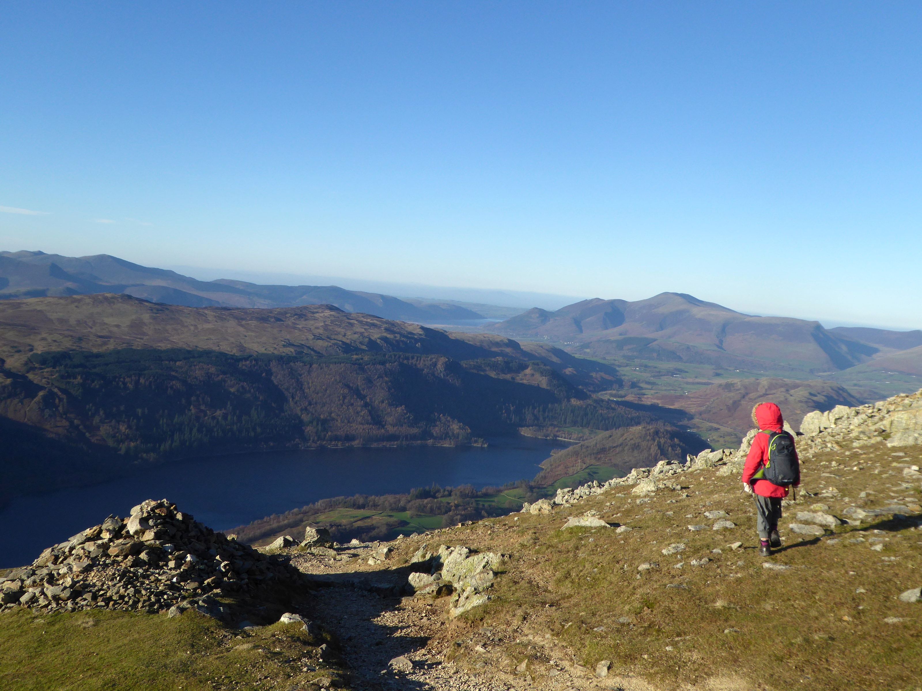 A walker in a red jacket picking along a rocky edge high above Thirlmere