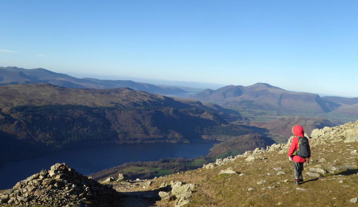 A walker in a red jacket picking along a rocky edge high above Thirlmere
