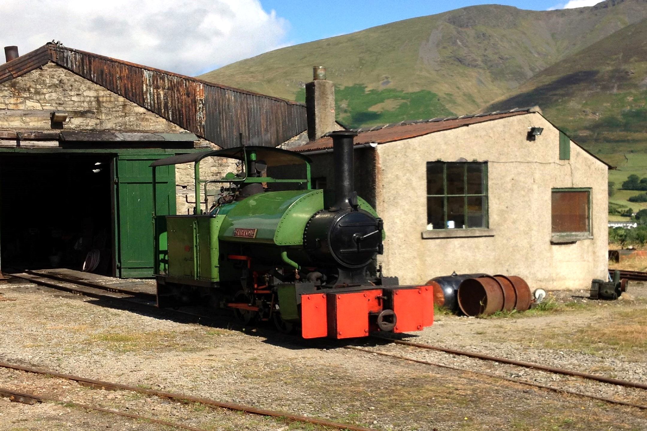 A small green steam locomotive standing outside the engine shed at the quarry museum