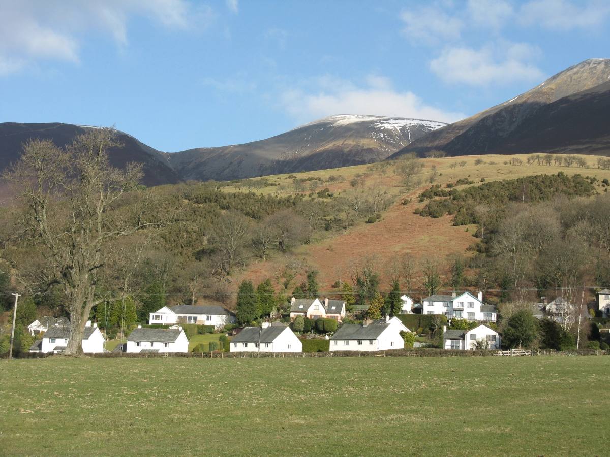 A row of whitewashed cottages in fields below fells with snow on the tops