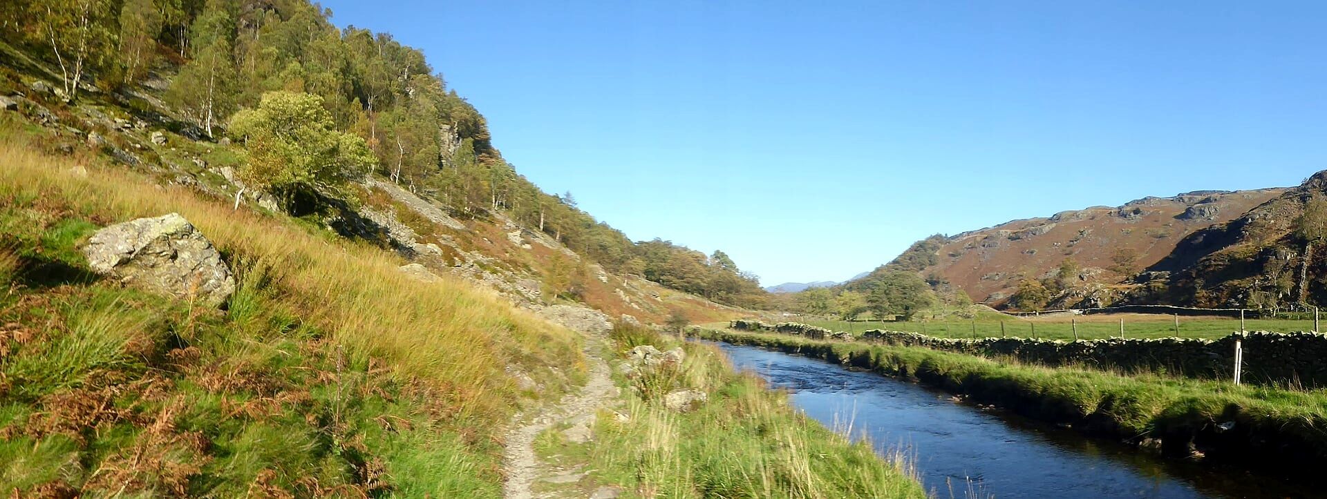 A path running along the riverbank below a steep grassy fellside