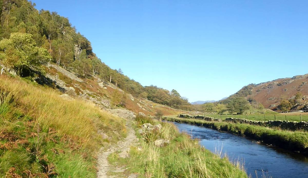A path running along the riverbank below a steep grassy fellside