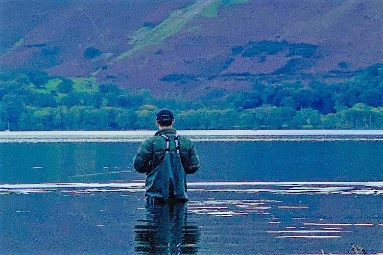 A paddleboarder standing out on flat calm water below the wooded fells