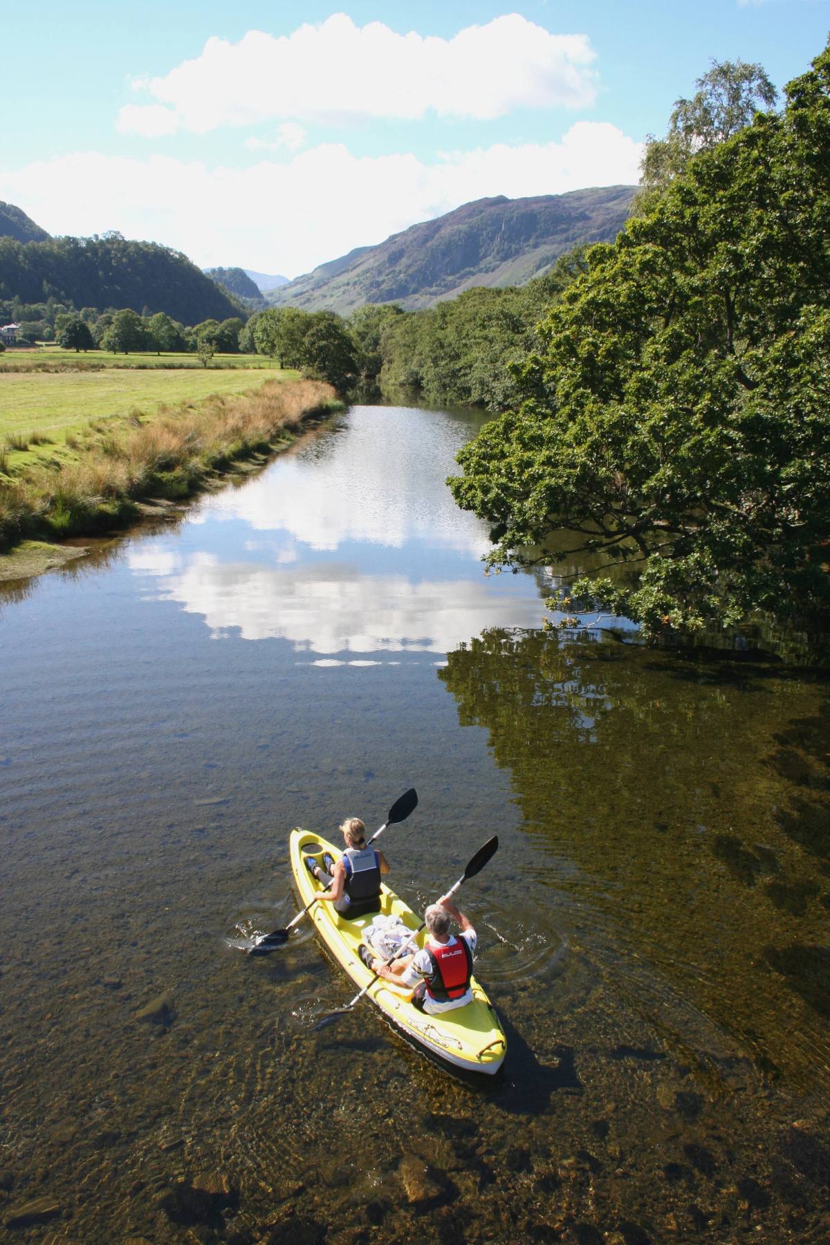 Kayakers on a still, tree-lined river reflecting the clouds