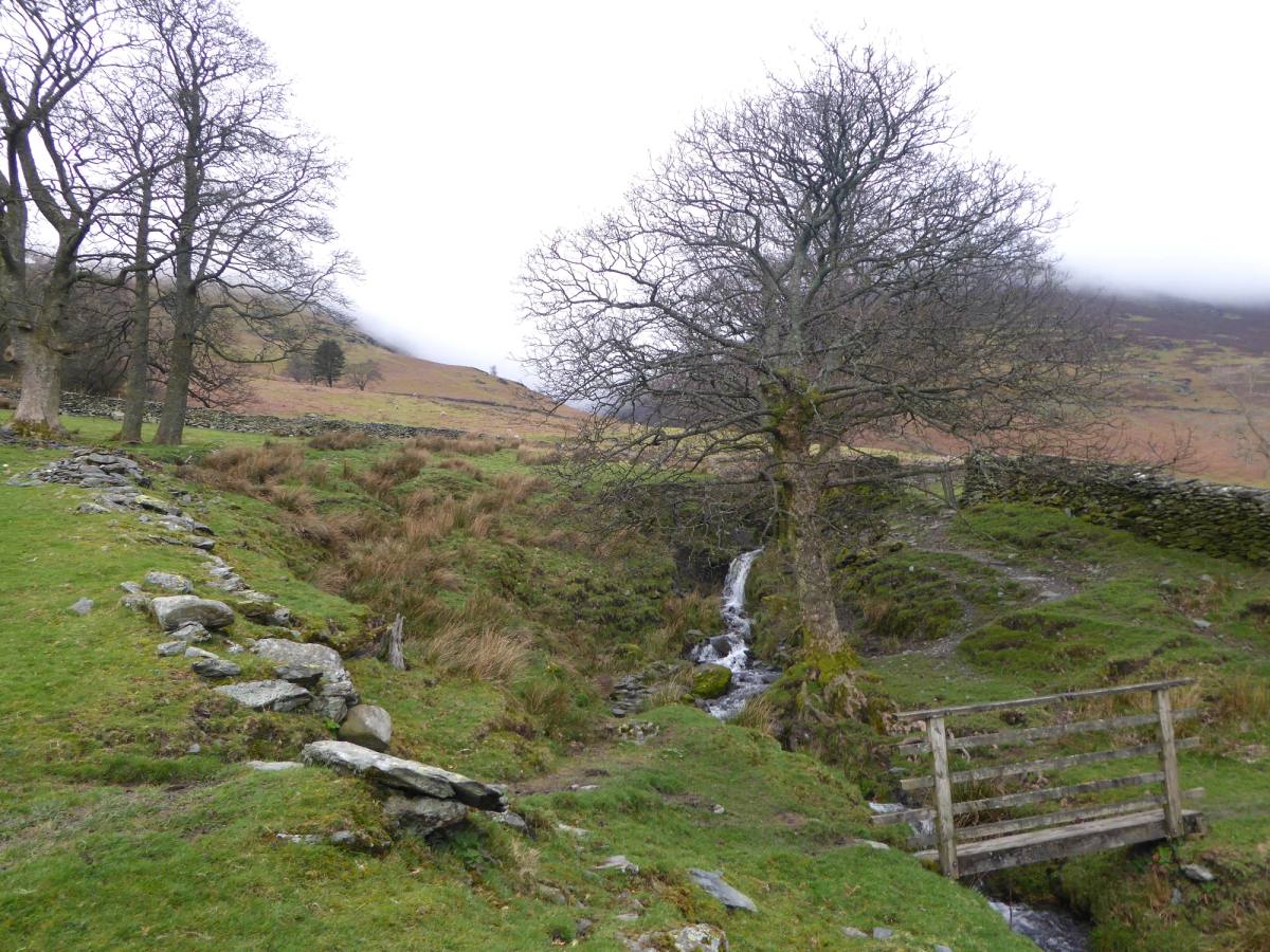 A wooden stile below a thin waterfall running down a rough grassy fellside