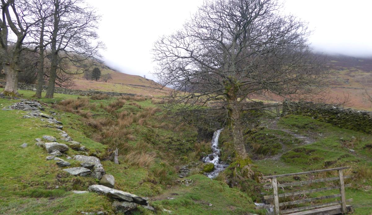 A wooden stile below a thin waterfall running down a rough grassy fellside