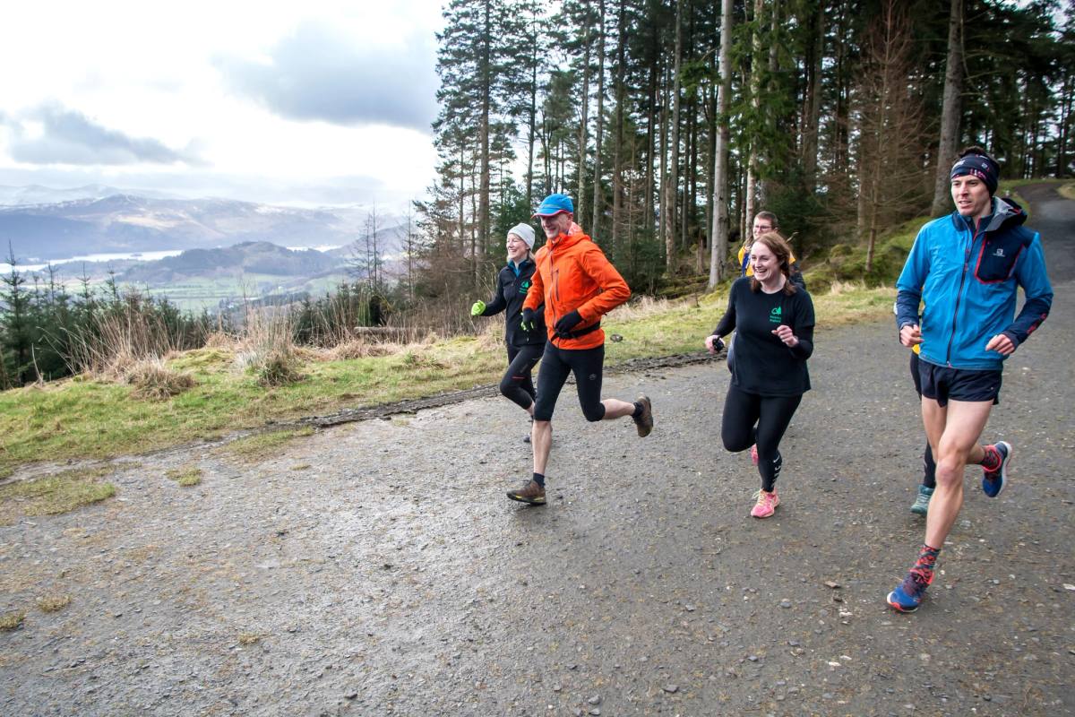 Three runners in a line coming down a broad forest road with the fells behind