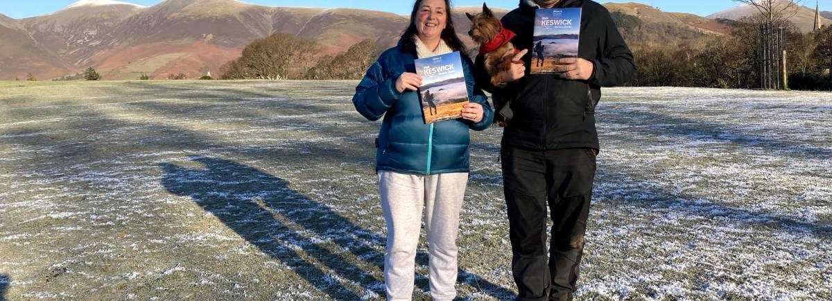 Vanessa Metcalfe and Rob Knowles standing in Crow Park with Skiddaw and Latrigg behind holding the Visit Keswick Guide. Robin is also holding his dog, Rosie