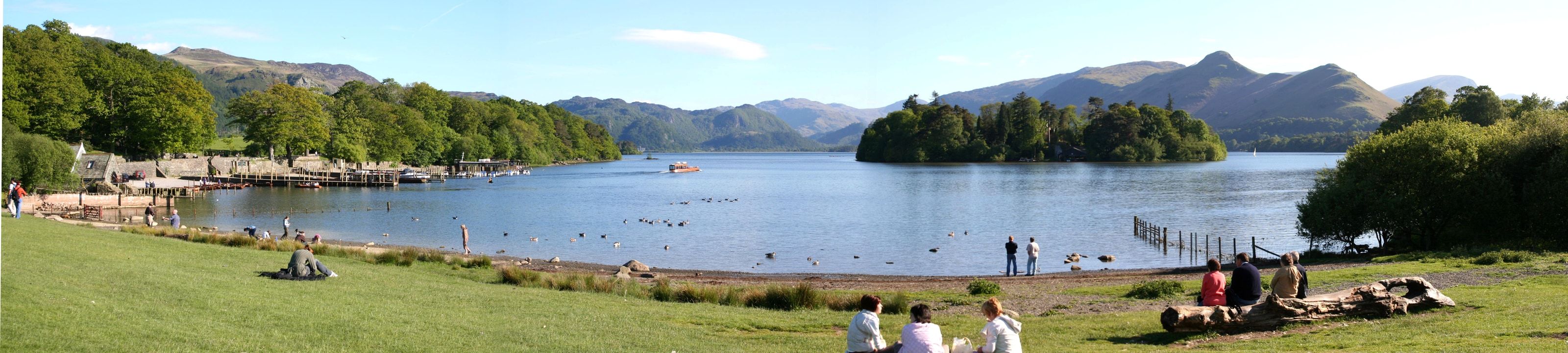 People sitting out on the grass along the Derwentwater shore with the islands beyond