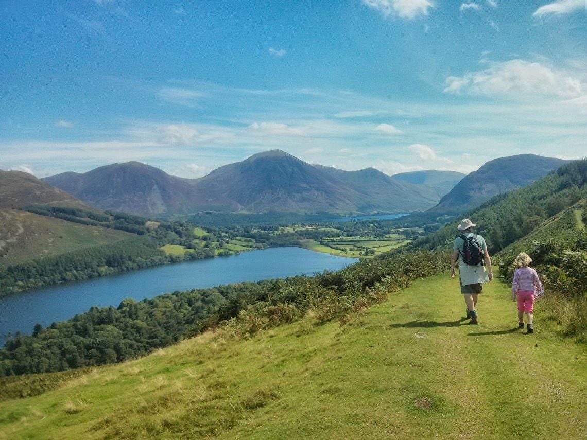 An adult and child walking a grassy path down towards Loweswater and the fells