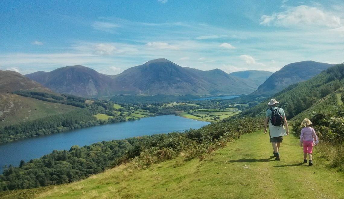 An adult and child walking a grassy path down towards Loweswater and the fells