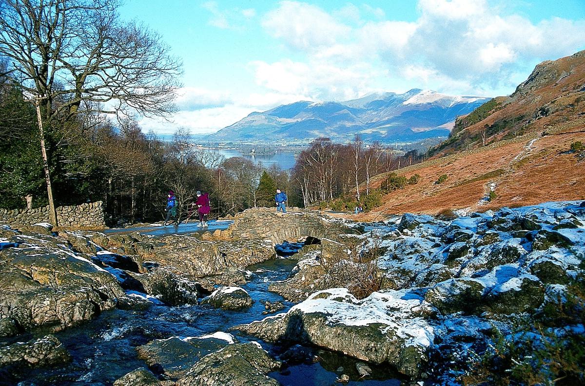 Walkers on Ashness Bridge above the tumbling beck with Skiddaw across the lake