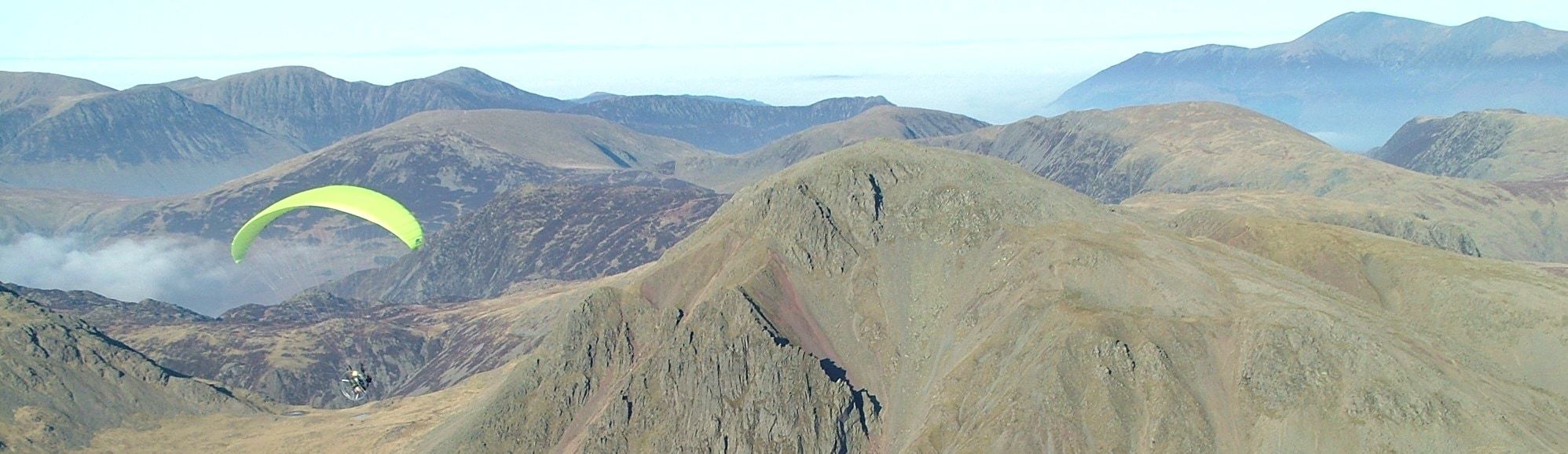 A paraglider turning above a broad grassy ridge with the fells beyond