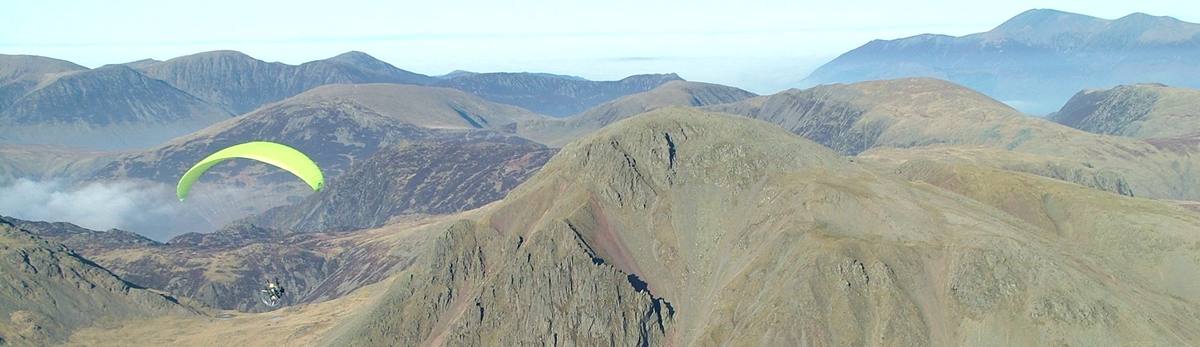A paraglider turning above a broad grassy ridge with the fells beyond