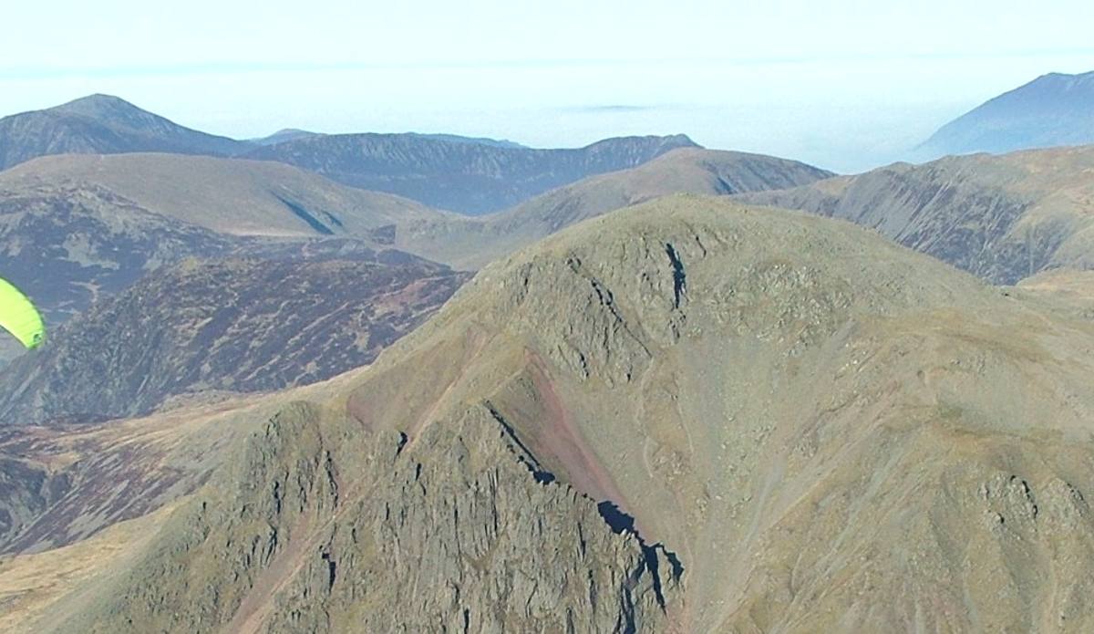 A paraglider turning above a broad grassy ridge with the fells beyond