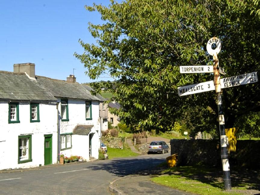 A white fingerpost at a village road junction beside a green-doored stone cottage