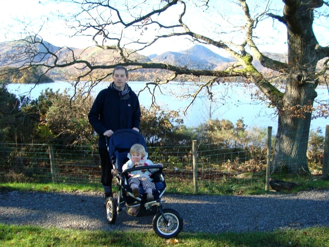 A man pushing a child in a buggy along a smooth track between old trees with the fells beyond
