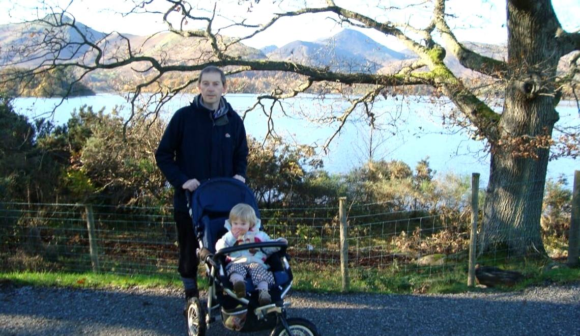 A man pushing a child in a buggy along a smooth track between old trees with the fells beyond