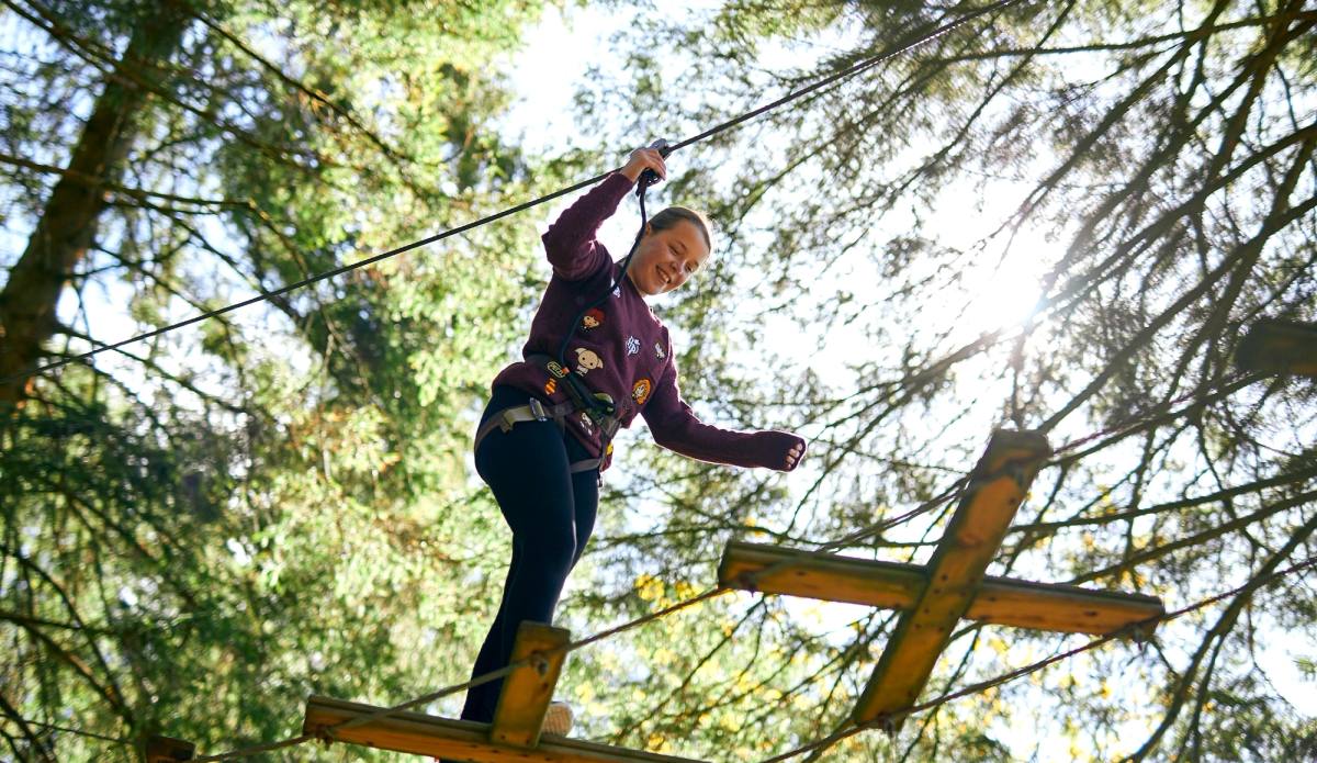 A person balancing along a high rope bridge in the tree canopy of an adventure course