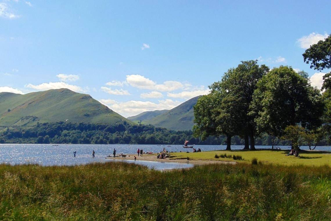 People on the grassy lake shore under mature trees with the fells opposite