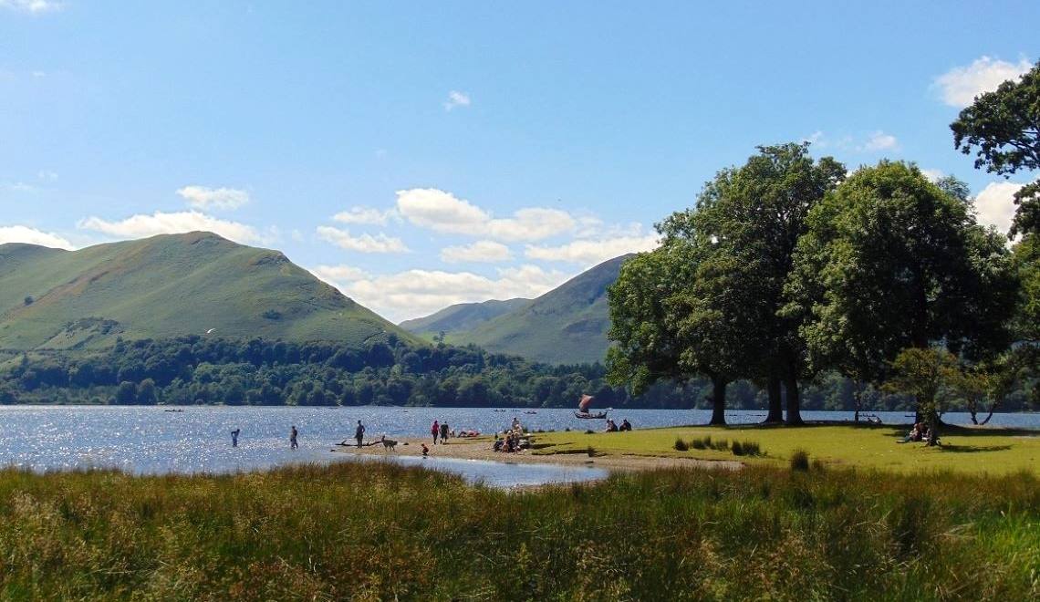 People on the grassy lake shore under mature trees with the fells opposite