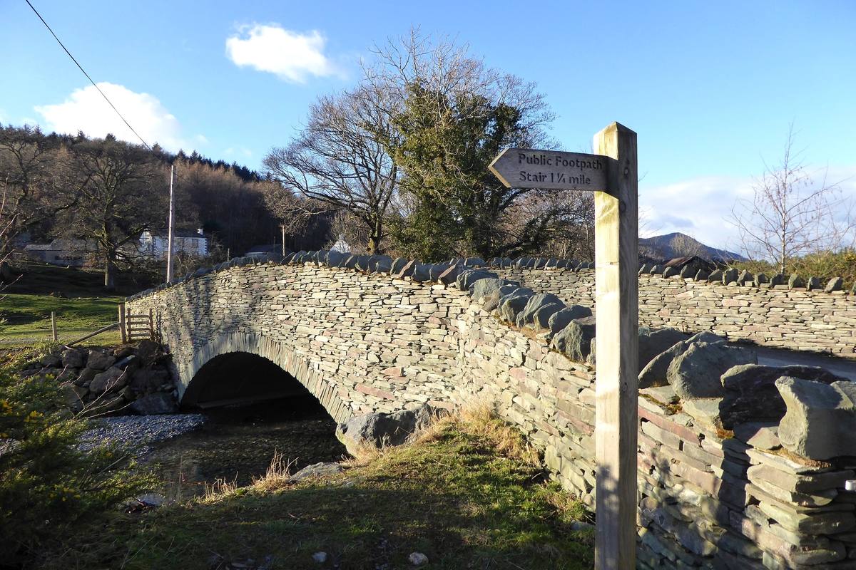 An old stone packhorse bridge over a beck with a wooden signpost beside it