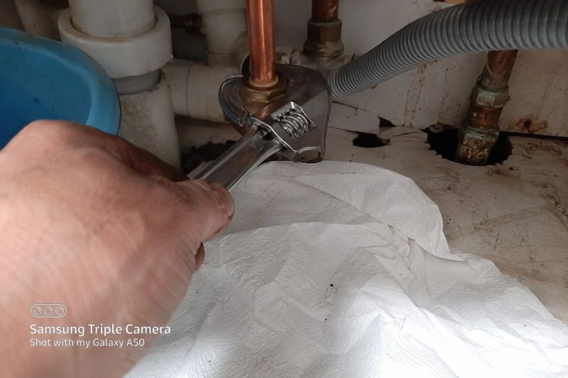 Engineer tightening a compression pipe fitting under a kitchen sink, paper towel laid underneath to check for drips
